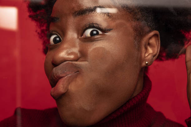 Close-up face of funny dark skinned young girl crushed on glass isolated on dark red studio background. Close-up face of funny dark skinned young girl crushed on glass isolated on dark red studio background. Concept of human emotions, facial expression, youth. Model leaning against glass crushed stock pictures, royalty-free photos & images