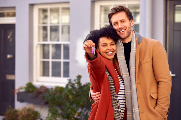 portrait d’un couple multiculturel à l’extérieur le jour du déménagement tenant les clés d’une nouvelle maison en automne ou en hiver - changement de logement photos et images de collection