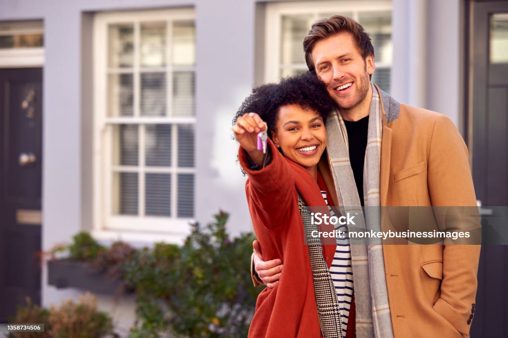 Portrait Of Multi Cultural Couple Outdoors On Moving Day Holding Keys To New Home In Fall Or Winter Home Ownership Stock Photo