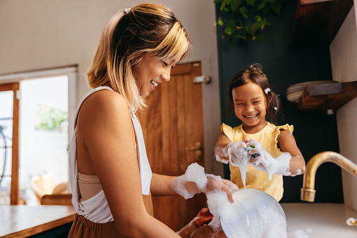 Happy mother and daughter washing dishes with bubbles. Mother and daughter having fun together in the kitchen. Loving single mother spending quality time with her young daughter at home.