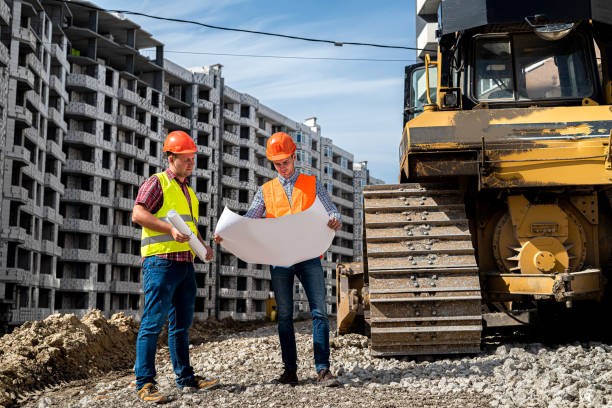 istock two young strong workers in uniform and helmets stand near a grader on a construction site 1358738560