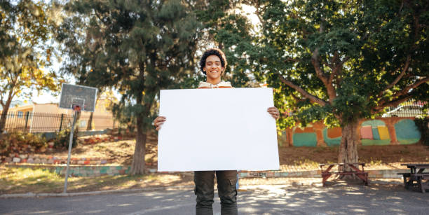 fröhlicher teenager mit einem leeren plakat in einem stadtpark - man holding a sign stock-fotos und bilder