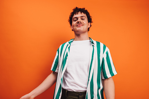 Young queer man looking at the camera with a cheerful face in a studio. Happy young generation z hipster smiling while standing alone against an orange background.
