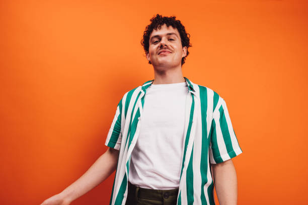 Young queer man looking at the camera cheerfully Young queer man looking at the camera with a cheerful face in a studio. Happy young generation z hipster smiling while standing alone against an orange background. queer man facing camera stock pictures, royalty-free photos & images