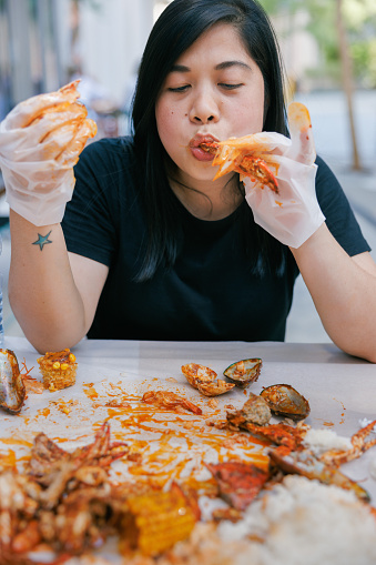 Woman sitting in sidewalk restaurant, eating traditional Dampa meal - seafood and white rice served spilled directly on the table with mussels, squid rings, crab and shrimps, wearing gloves and sucking crab