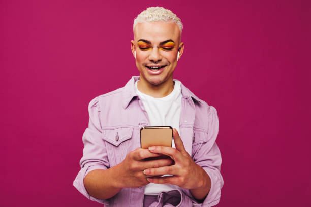 Queer man playing his favourite playlist Queer man playing his favourite playlist. Happy young man using a smartphone while wearing wireless earphones in a studio. Non-conforming queer man smiling cheerfully against a purple background. generation z stock pictures, royalty-free photos & images