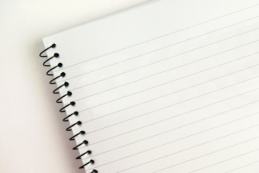 Horizontal picture of a white page of a single lined spiral bound notepad. The punch holes of the note pad are round and the spiral wire is black in colour. There is a blank plain paper sheet below by the notepad. There is no text, no people and ample copy space.