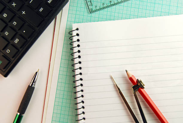 istock Geometry tools - An open compass placed over a page of a spiral notepad and graph paper, with calculator and set square in the hindsight. 1358743622