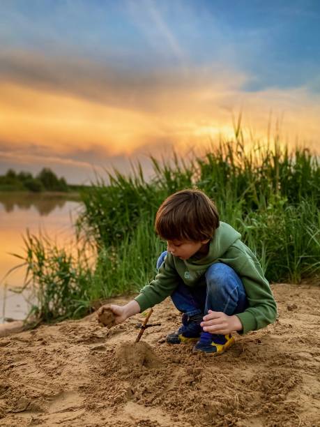 Blonde boy is making his sand castle at the edge of the lake, orange sunset sky background, green grass at right, green hoodie, childhood, playing games, summertime