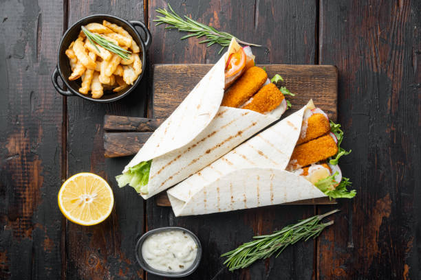 Sandwich roll with fish fingers, cheese and vegetables, on wooden cutting board, on old dark  wooden table background, top view flat lay Sandwich roll with fish fingers, cheese and vegetables set, on wooden cutting board, on old dark  wooden table background, top view flat lay fish stick stock pictures, royalty-free photos & images