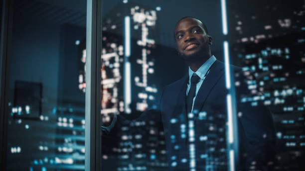 Successful Black Businessman in a Tailored Suit Standing in His Office Looking out of the Window on Night City. Successful Investment Manager Working Late Planning e-Commerce Service Purchase Strategy Successful Black Businessman in a Tailored Suit Standing in His Office Looking out of the Window on Night City. Successful Investment Manager Working Late Planning e-Commerce Service Purchase Strategy passion stock pictures, royalty-free photos & images