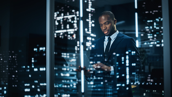 Portrait of Stylish Black Businessman Using Laptop, Looking out of Window on a City. successful African-American CEO Working on Computer, Working Hard Late At Night to Achieve Best Results