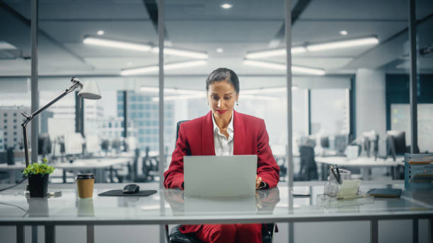 exitosa mujer de negocios caucásica con traje perfectamente elegante que trabaja en una computadora portátil en la oficina del piso superior con vistas a la gran ciudad. mujer ceo gerente de la empresa. toma de vista frontal - fortaleza estructura de edificio fotografías e imágenes de stock