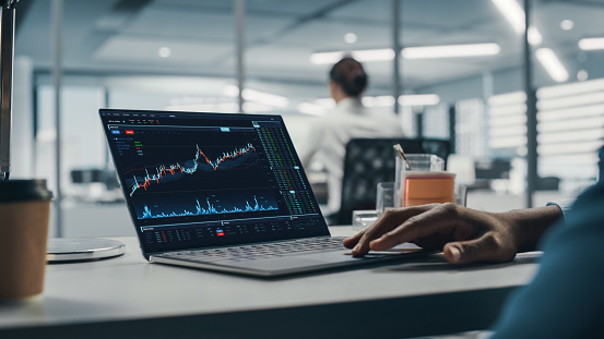 Trader holding cup of coffee and text messaging on smart phone while sitting in front of stock market reports on computer screens at workplace