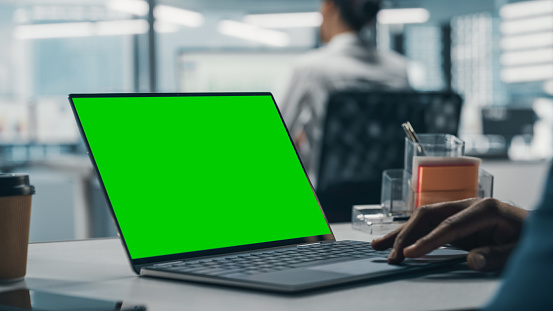Successful Black Businessman Sitting at Desk Working on Green Screen Laptop in Office. African American Businessperson using Touch Pad on Chroma Key Computer. Shot with Focus on Hand