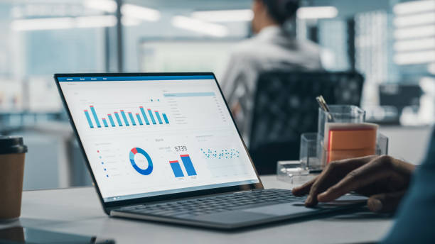 exitoso hombre de negocios negro sentado en el escritorio trabajando en una computadora portátil en la oficina de la gran ciudad. top manager trabajando duro haciendo investigación para el proyecto de comercio electrónico con graps, gráficos y tablas.  - sheet fotografías e imágenes de stock