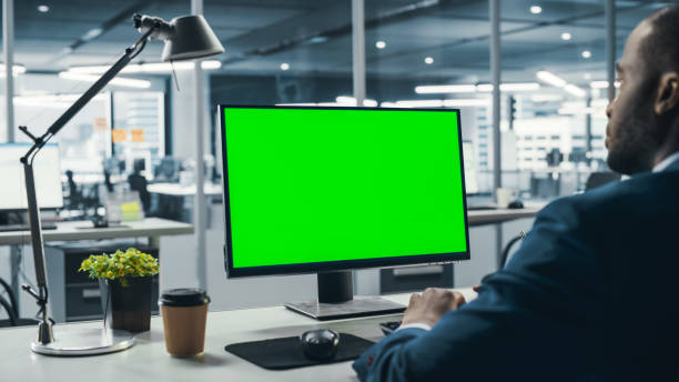istock Successful Black Businessman Sitting at Desk Working on Green Screen Laptop Computer in Office. African American Businessperson using Chroma Key Display. Over the Shoulder Shot 1358748579