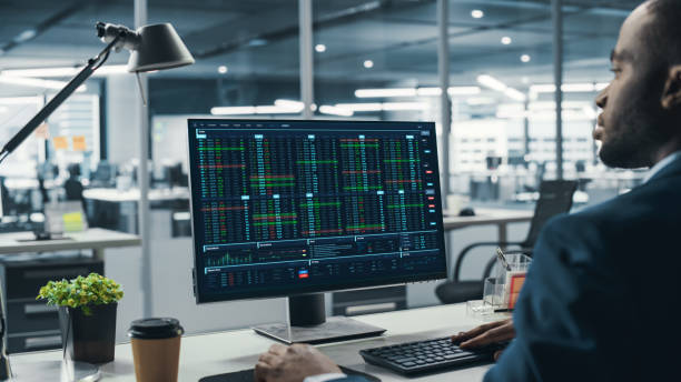 istock Successful Black Businessman Sitting at Desk Working on Desktop Computer in Big City Office. Hard Working Top Manager Doing Research for e-Commerce Project with Graps, Charts and Tables. Over Shoulder 1358748582