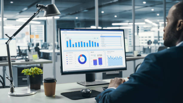 istock Successful Black Businessman Sitting at Desk Working on Desktop Computer in Big City Office. Hard Working Top Manager Doing Research for e-Commerce Project with Graps, Charts and Tables. Over Shoulder 1358748587
