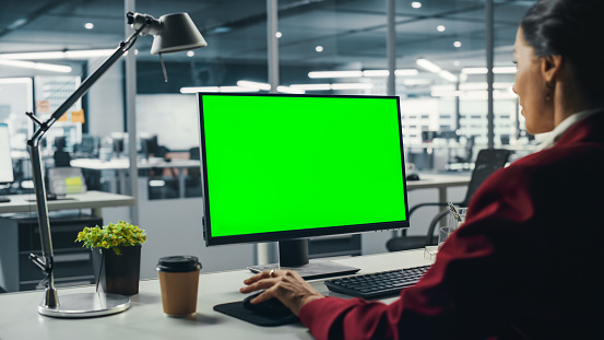 Successful Caucasian Businesswoman Sitting at Desk Working on Green Screen Laptop Computer in Office. Beautiful Businessperson using Chroma Key Display. Over the Shoulder Shot