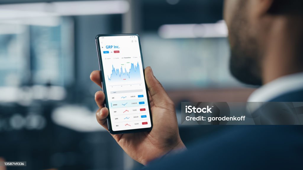 Black Businessman Holding Smartphone and Checking Stock and Cryptocurrency Market in Office. African-American Businessperson using Internet with Mobile Phone Device. Over Shoulder Shot Telephone Stock Photo