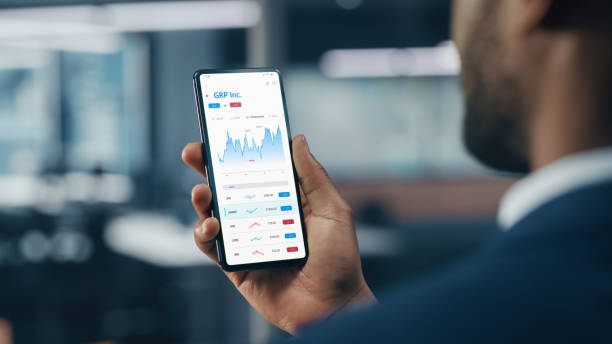 Black Businessman Holding Smartphone and Checking Stock and Cryptocurrency Market in Office. African-American Businessperson using Internet with Mobile Phone Device. Over Shoulder Shot Black Businessman Holding Smartphone and Checking Stock and Cryptocurrency Market in Office. African-American Businessperson using Internet with Mobile Phone Device. Over Shoulder Shot man looking at phone over shoulder stock pictures, royalty-free photos & images