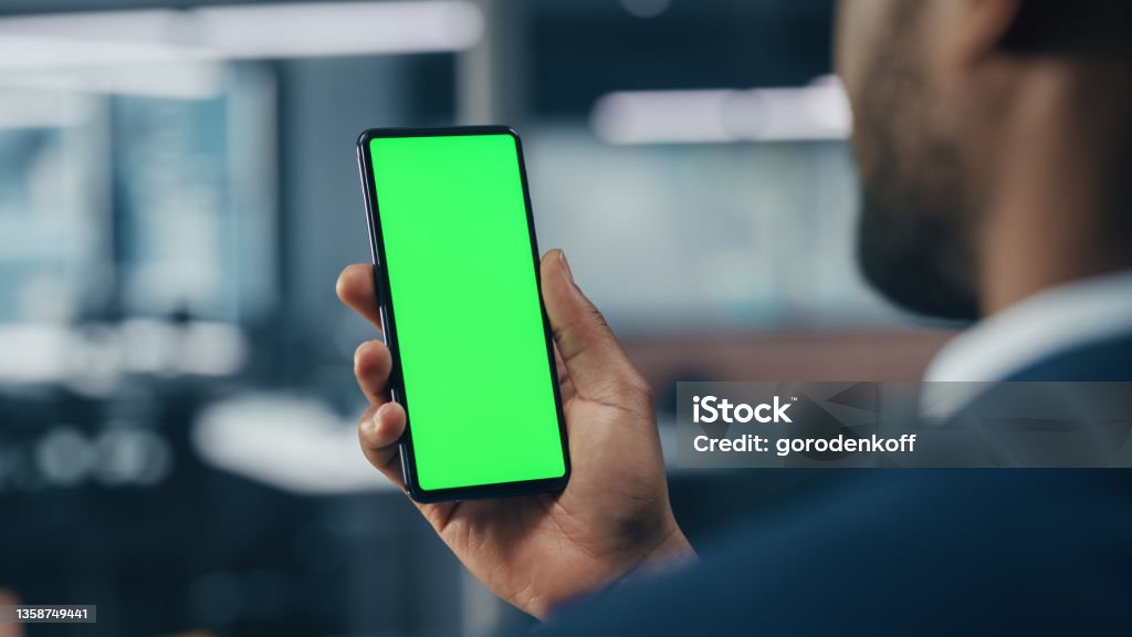 Black Businessman with Green Screen Chroma Key Smartphone in Office. African-American Businessperson using Internet, Social Media, Online Shopping with Mobile Phone Device. Over Shoulder Chroma Key Stock Photo