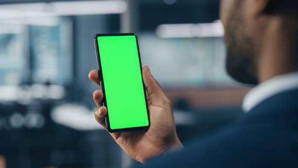 Black Businessman with Green Screen Chroma Key Smartphone in Office. African-American Businessperson using Internet, Social Media, Online Shopping with Mobile Phone Device. Over Shoulder Black Businessman with Green Screen Chroma Key Smartphone in Office. African-American Businessperson using Internet, Social Media, Online Shopping with Mobile Phone Device. Over Shoulder personal perspective standing stock pictures, royalty-free photos & images