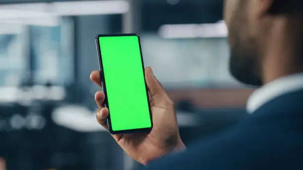 Black Businessman with Green Screen Chroma Key Smartphone in Office. African-American Businessperson using Internet, Social Media, Online Shopping with Mobile Phone Device. Over Shoulder