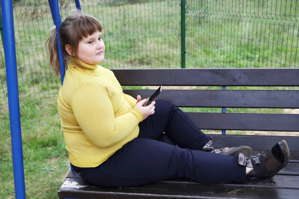 plumpes nachdenkliches mädchen in gelbem pullover sitzt allein auf der schaukelbank im freien, hält smartphone - child obesity stock-fotos und bilder