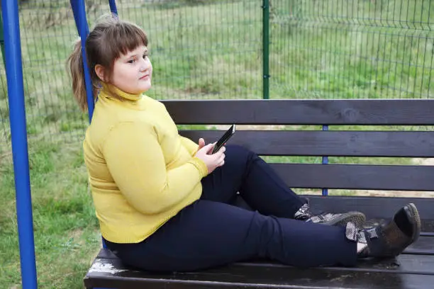 Photo of Plump thoughtful girl in yellow sweater sits alone on bench of swing outdoors, hold smartphone