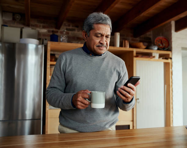 multi-cultural elderly male reading daily news on smartphone while standing in modern kitchen - senior adult human face male action imagens e fotografias de stock