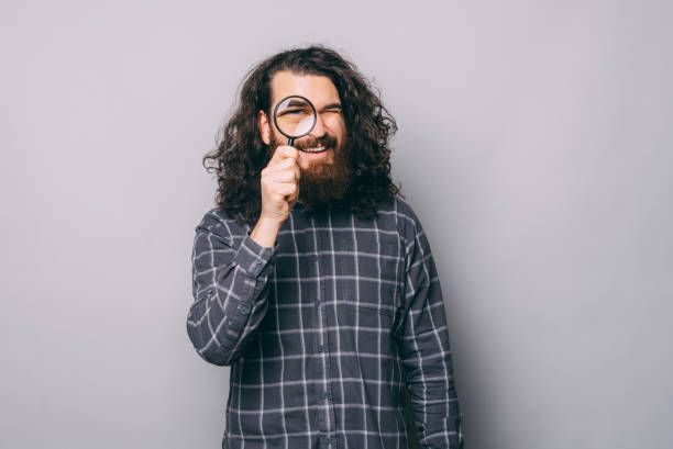 Portrait of young bearded man looking trough magnifying glass over grey background stock photo