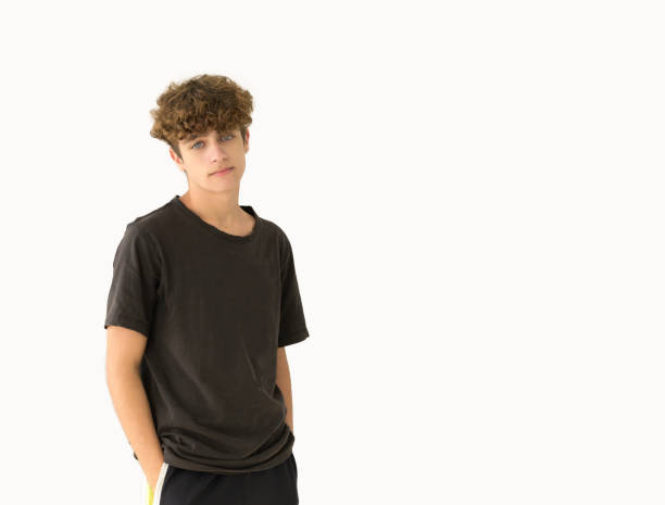 Young teenage boy in black t-shirt looking at camera on white background. Young man in black tshirt and shorts standing on white background blank expression stock pictures, royalty-free photos & images