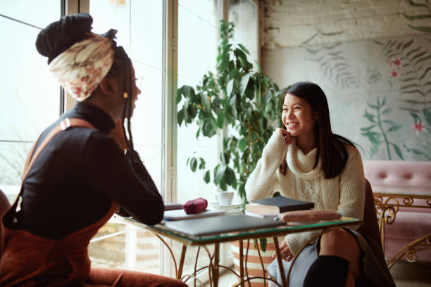 Due felici ragazze universitarie multiculturali siedono nella caffetteria, chiacchierando e spettegolando. Studentesse e ragazze parlano. - foto stock