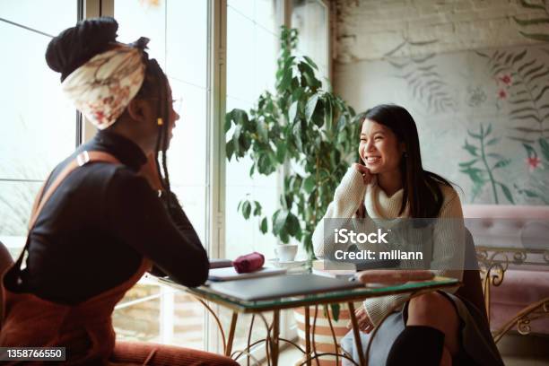 Two Happy Multicultural Female College Girls Sit In The Cafeteria Chatting And Gossiping Female Students And Girls Talk Stock Photo - Download Image Now