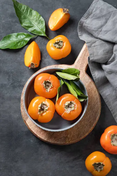 Fresh ripe persimmon fruits in bowl on a cutting board on a dark background with slices and leaves. The concept of healthy nutrition and vitamins. Top view.