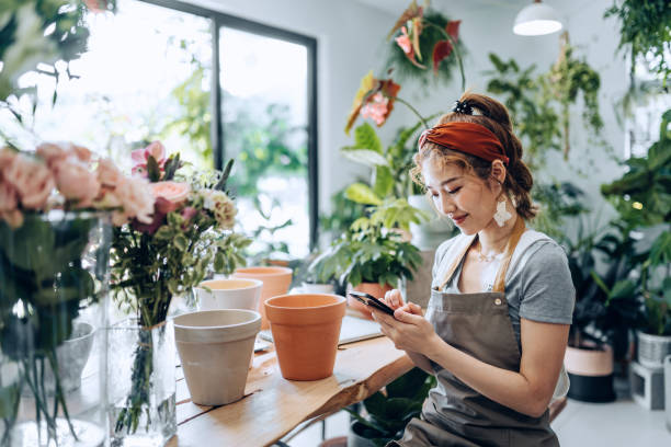 giovane fiorista asiatica, proprietaria di un negozio di fiori di piccole imprese, che utilizza lo smartphone al bancone del negozio contro fiori e piante. controllo delle scorte, ricezione degli ordini dei clienti, vendita di prodotti online. routine quot - florist telephone flower business foto e immagini stock