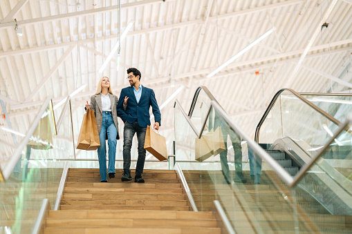 Low-angle view of happy beautiful young couple holding shopping paper bags with purchases and walking down stairs, holding hands at mall. Concept of holiday sales, retail, consumerism.