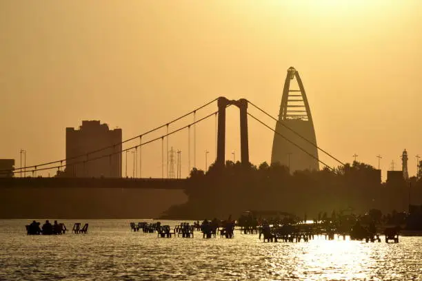 Photo of Suset scene at Sudanese capital Khartoum with people sitting on plastic chairs in the shallow Nile riverside.