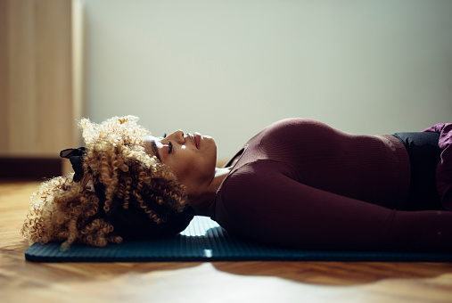 Side view of an Afro-American woman lying on the yoga mat on the floor. She might be doing breathing exercises. She is in a fitness studio or in her room during the day and she might be a yoga student or an instructor.