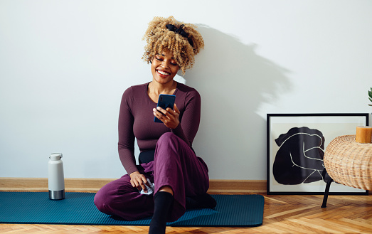 Afro-American woman is sitting on the yoga mat on the floor and looking at her phone. She looks happy and a bit tired. She might be taking a break during an exercise session. She is in a fitness studio or in her room during the day and she might be a yoga student or an instructor.