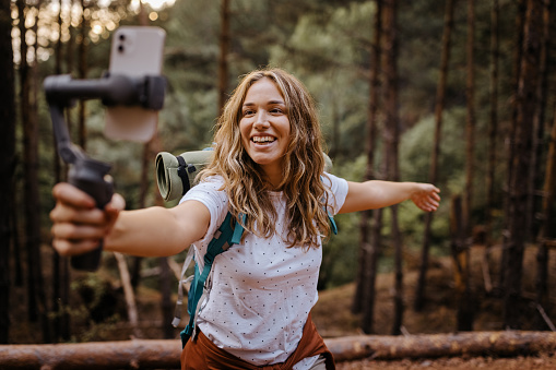 Cheerful young female vlogger with backpack during hiking adventure recording vlog and going live for her audience on social media