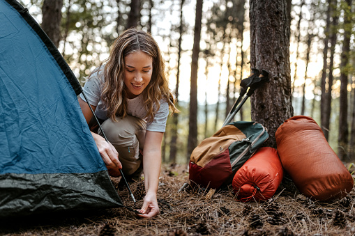 Beautiful smiling women setting up tent in forest with backpack resting near tree