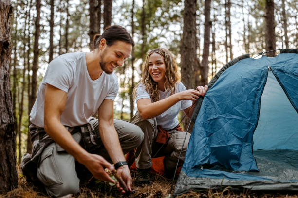 Couple building tent in forest during hike Beautiful smiling women with handsome boyfriend setting up tent in forest while talking camping stock pictures, royalty-free photos & images