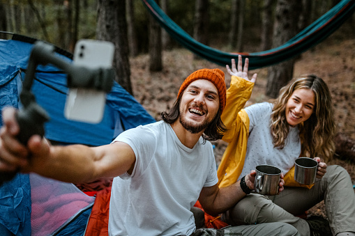 Smiling young man and woman enjoying hot coffee and doing video call and recording vlog using smartphone sitting outside tent during hiking