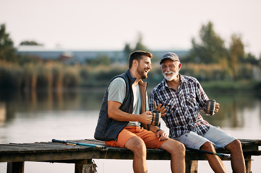 Happy senior fisherman and his adult son talking while having a drink on a pier. Copy space.
