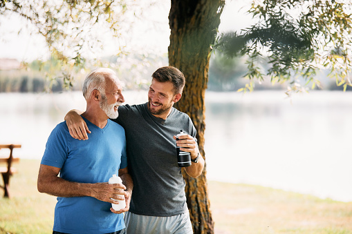 Happy athletic man and his mature father talking while drinking water and walking after sports training in nature. Copy space.