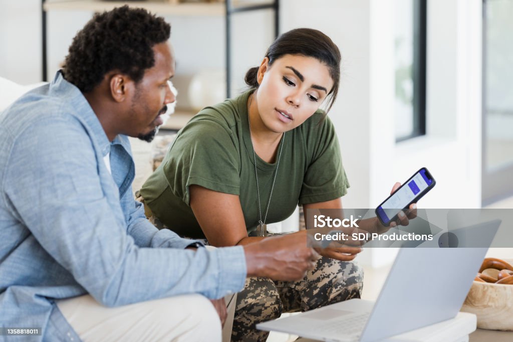 Man and soldier wife discuss finances using phone and laptop The mid adult man holds a credit card as he and his soldier wife discuss home finances.  Her smart phone has an app displayed on the screen. Military Stock Photo