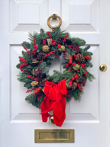Christmas mood: festive christmasy themed winter natural wreath on a white wooden door. Decorated with red tied bow, berries, pine cones and fir tree